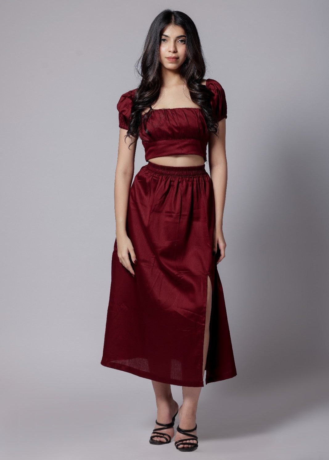 A model wearing a wine-colored skirt set with a crop top and a long skirt with a side slit, with sleek neckline and chic fit.