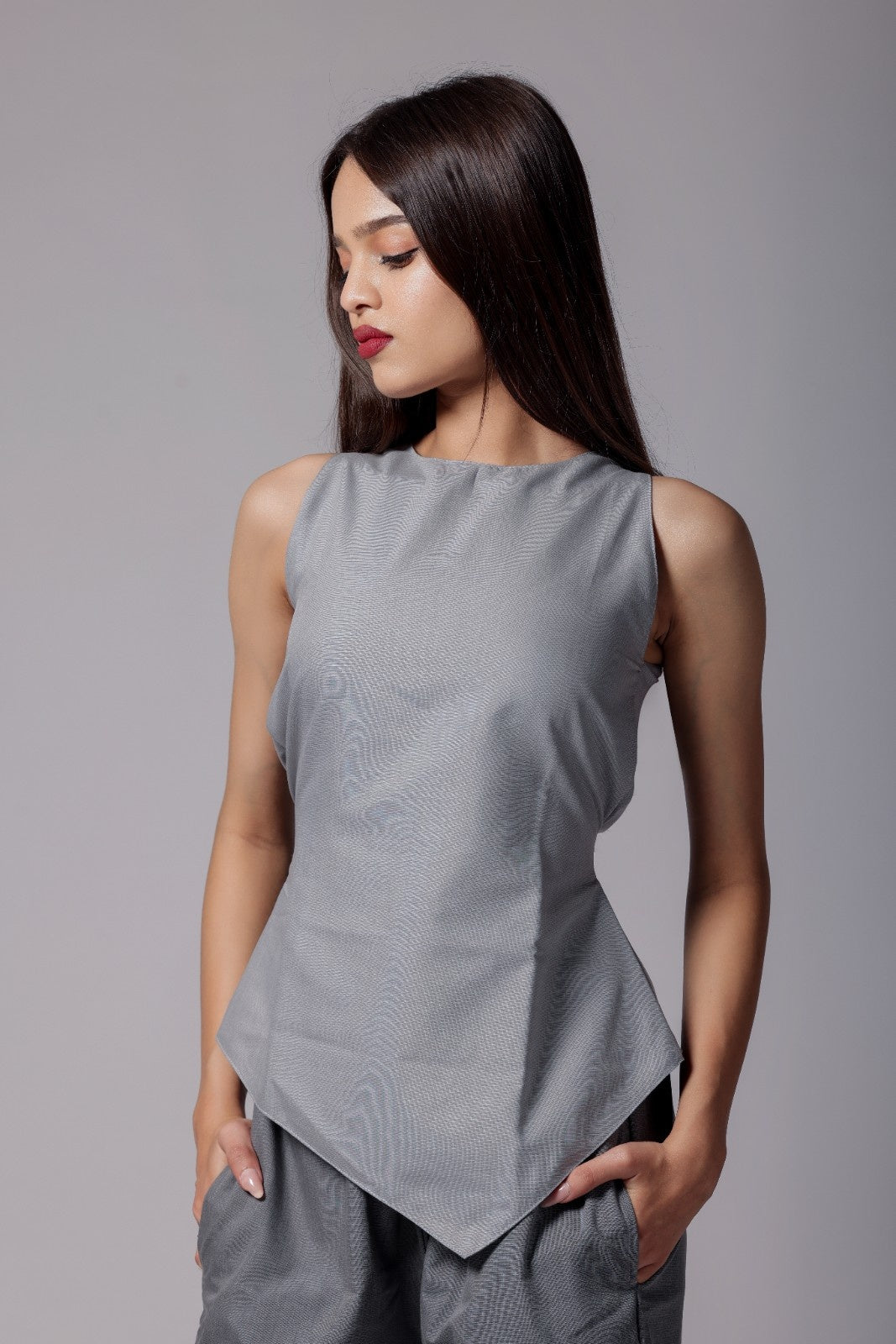 A woman wearing terry cotton co-ord set with an asymmetrical sleeveless sleek top in light gray with back tie up bow and tailored dark gray pants with pockets.