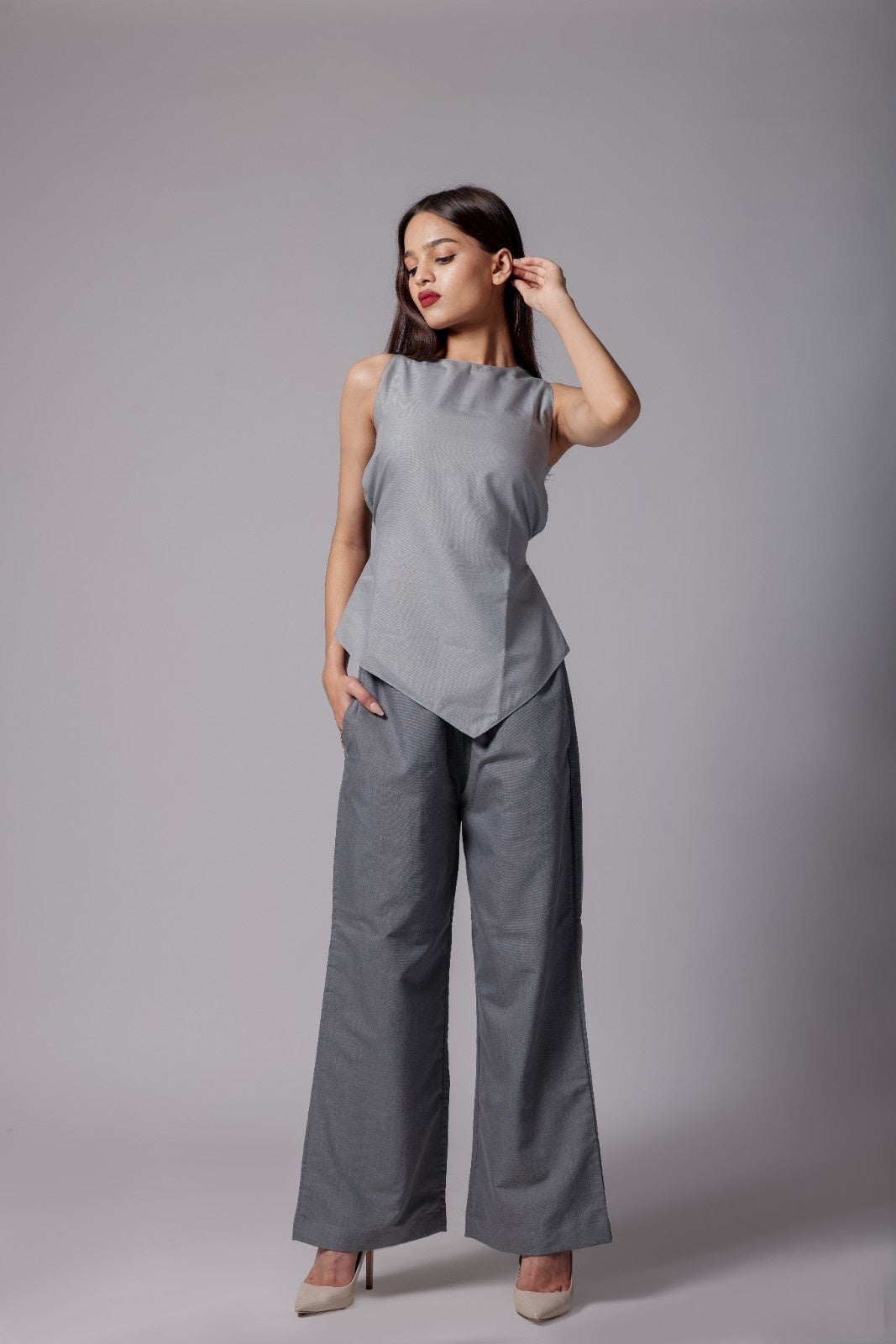 A woman wearing terry cotton co-ord set with an asymmetrical sleeveless sleek top in light gray with back tie up bow and tailored dark gray pants with pockets.