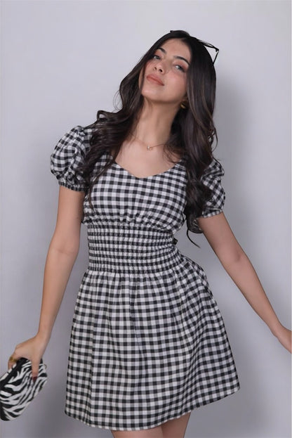 A woman wearing gingham check-patterned, V-neckline, puff sleeve, ruched waist flared, short dress with an elastic waist.