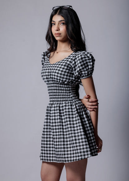 A woman wearing gingham check-patterned, V-neckline, puff sleeve, ruched waist flared, short dress with an elastic waist.