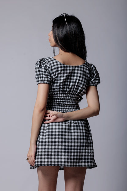 A woman wearing gingham check-patterned, V-neckline, puff sleeve, ruched waist flared, short dress with an elastic waist.