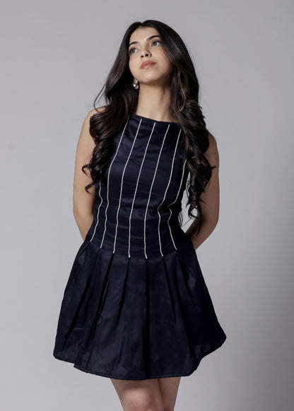 A woman in a navy blue short dress with white piping, a high neck, and sleeveless design. The dress has pleats and a flare at the lower part and features a bow at the back.