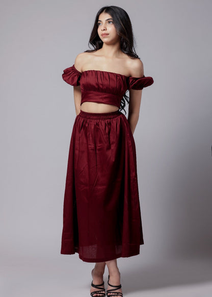 A model wearing a wine-colored skirt set with a crop top and a long skirt with a side slit, with sleek neckline and chic fit. off shoulder top