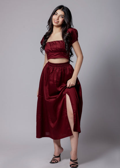 A model wearing a wine-colored skirt set with a crop top and a long skirt with a side slit, with sleek neckline and chic fit.