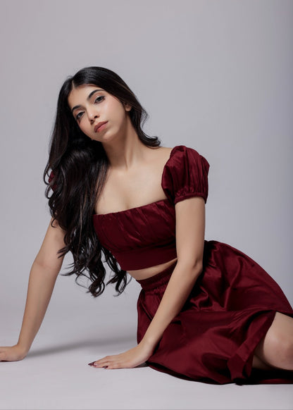 A model wearing a wine-colored skirt set with a crop top and a long skirt with a side slit, with sleek neckline and chic fit.