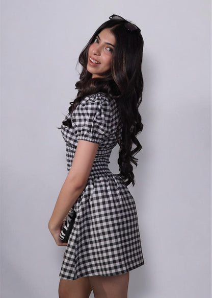 A woman wearing gingham check-patterned, V-neckline, puff sleeve, ruched waist flared, short dress with an elastic waist.
