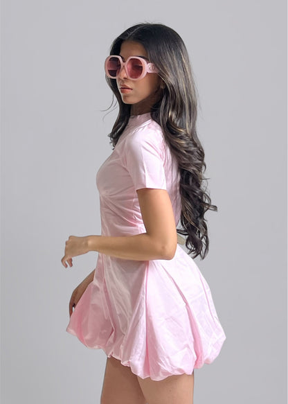 A woman wearing a bubblegum pink dress with short sleeves and a round neckline, paired with sunglasses.